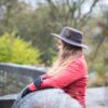 Halter-Highclere-scaled-2.jpg A woman with long hair in a grey felt hat decorated with a feather and a red quilted jacket, wearing black gloves, leans on a wet stone bridge and gazes across a rainy, blurred riverside woodland.