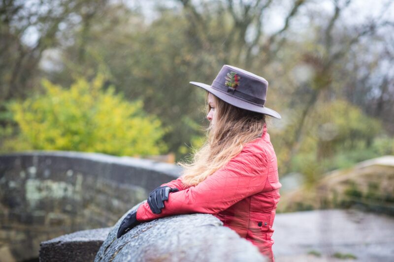 Halter-Highclere-scaled-2.jpg A woman with long hair in a grey felt hat decorated with a feather and a red quilted jacket, wearing black gloves, leans on a wet stone bridge and gazes across a rainy, blurred riverside woodland.