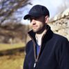 A man wearing a black baseball cap and dark zip-up coat with the collar turned up stands beside a sunlit stone wall, looking thoughtful against a blurred background of bare trees and a grassy field.