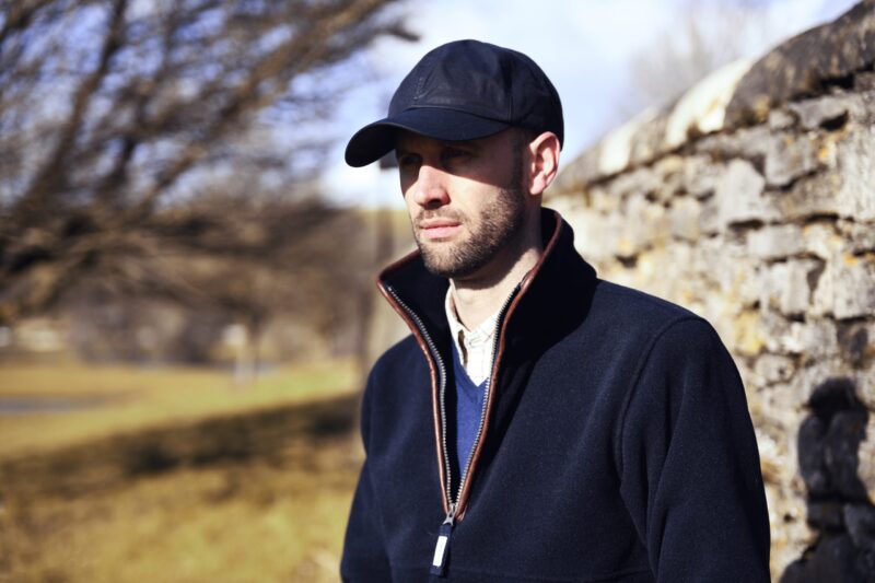 A man wearing a black baseball cap and dark zip-up coat with the collar turned up stands beside a sunlit stone wall, looking thoughtful against a blurred background of bare trees and a grassy field.