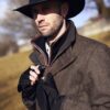 Hawk-and-Highclere-9-scaled-2.jpg Man wearing a wide-brimmed black hat with a feather and a brown herringbone coat, fastening black gloves while standing outdoors in bright sunlight with blurred trees and fields behind him.