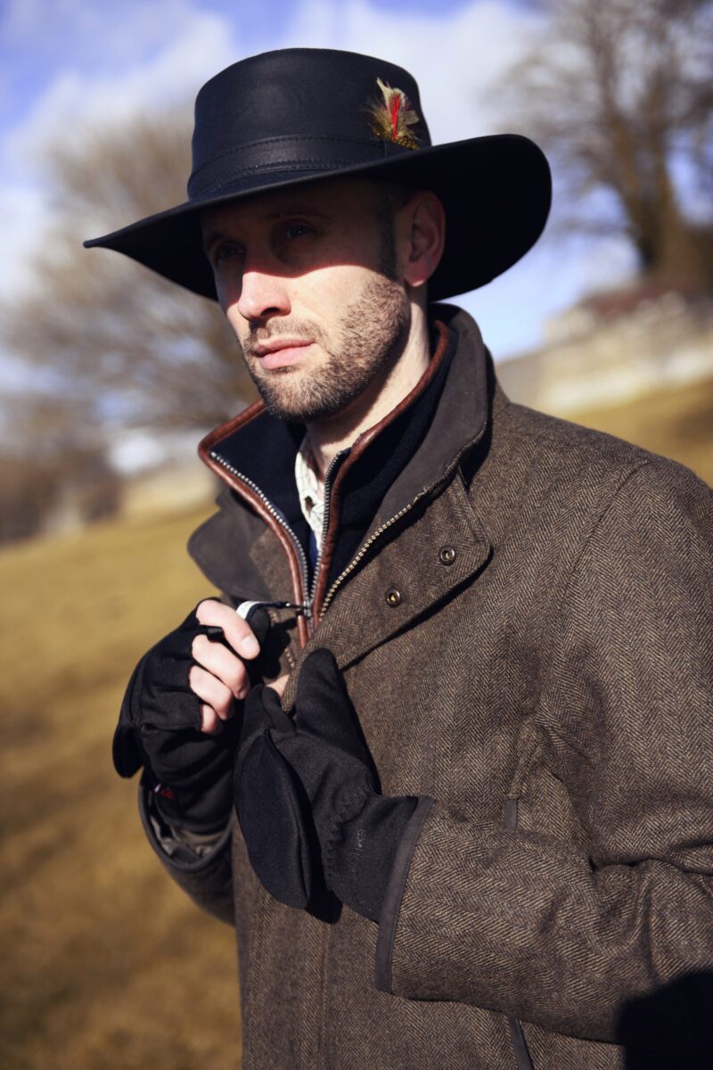 Hawk-and-Highclere-9-scaled-2.jpg Man wearing a wide-brimmed black hat with a feather and a brown herringbone coat, fastening black gloves while standing outdoors in bright sunlight with blurred trees and fields behind him.