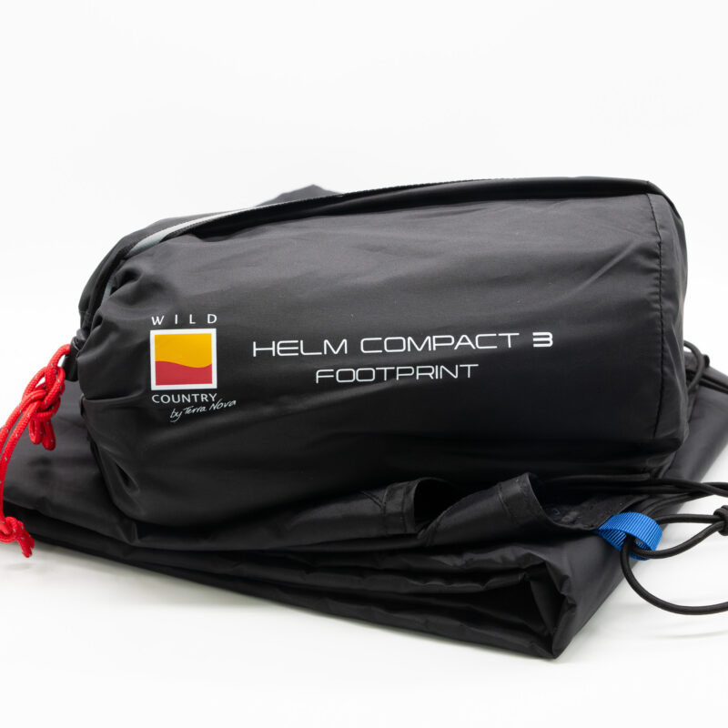 Black storage bag labeled HELM COMPACT 3 FOOTPRINT with Wild Country logo, red drawcord and blue tab, containing a folded tent footprint on a white background.