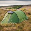 Hoolie-Compact-2-2-scaled-2.jpg Green tunnel tent secured with bright yellow guy lines pitched among tall grass and white cottongrass on a windswept moor, with a rocky escarpment and a band of trees beneath a cloudy sky in the background.
