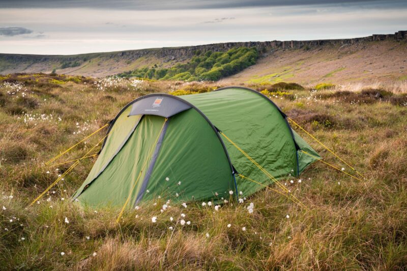 Hoolie-Compact-2-2-scaled-2.jpg Green tunnel tent secured with bright yellow guy lines pitched among tall grass and white cottongrass on a windswept moor, with a rocky escarpment and a band of trees beneath a cloudy sky in the background.