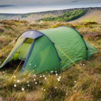 A green tunnel tent pegged with yellow guy lines pitched on windswept heather and cotton-grass moorland with low rocky cliffs and a cloudy sky in the background.