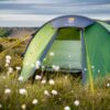 Terra Nova Shoot Green dome tent with its front door open and bright yellow guy lines, pitched in a grassy field studded with white cotton-like flowers and low cliffs under a cloudy sky.