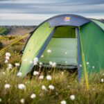 Green dome tent with its front door open and bright yellow guy lines, pitched in a grassy field studded with white cotton-like flowers and low cliffs under a cloudy sky.