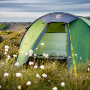 Green dome tent with its front door open and bright yellow guy lines, pitched in a grassy field studded with white cotton-like flowers and low cliffs under a cloudy sky.