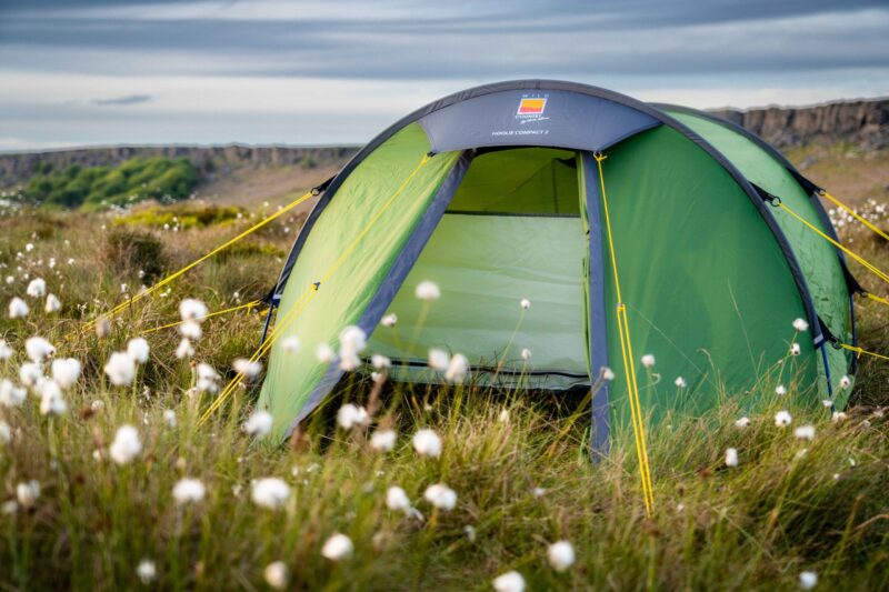 Terra Nova Shoot Green dome tent with its front door open and bright yellow guy lines, pitched in a grassy field studded with white cotton-like flowers and low cliffs under a cloudy sky.