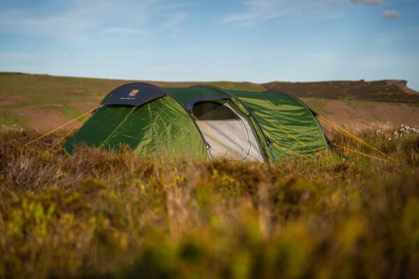 Hoolie-Compact-2-ETC-1-scaled-2.jpg A green tunnel-style camping tent with yellow guy lines and an open grey inner door pitched among grassy heather on rolling moorland beneath a blue sky.