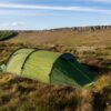 Terra Nova Shoot Low green tunnel tent pitched on a grassy upland moor among heather and white cotton grass, with a low rocky escarpment ridge in the background beneath a blue sky with thin clouds.