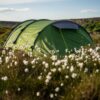 Terra Nova Shoot Green tunnel tent with yellow guy lines pitched on a windswept moor surrounded by white cotton‑grass flowers, with rolling cliffs and a pale blue sky in the background.