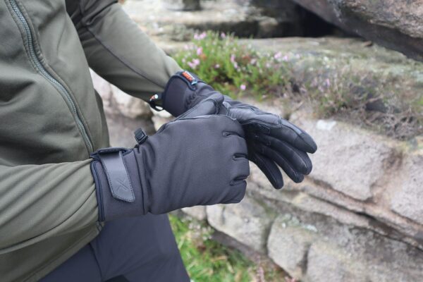 Person in an olive-green jacket putting on black insulated gloves with wrist straps and textured leather palms beside lichen-covered rocks and small pink flowers