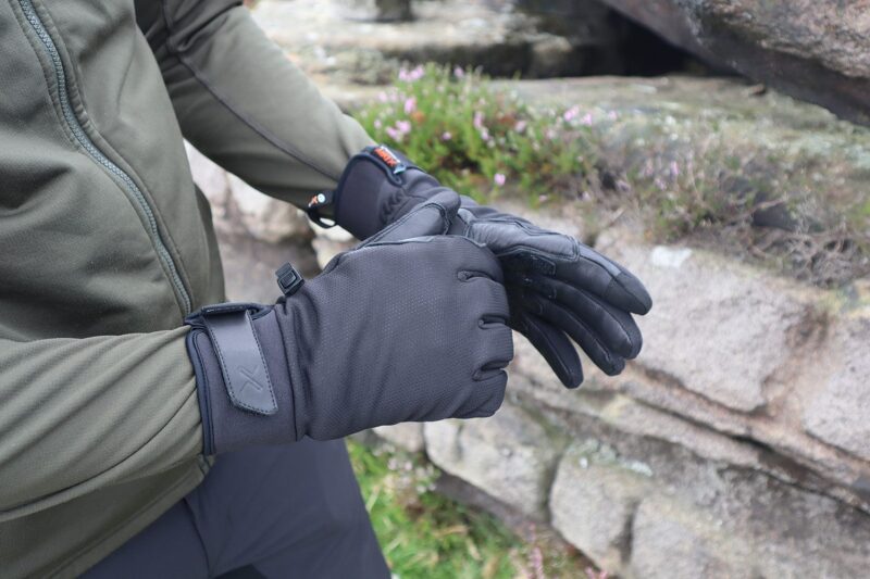 IMG_2168-3.jpg Person in an olive-green jacket putting on black insulated gloves with wrist straps and textured leather palms beside lichen-covered rocks and small pink flowers