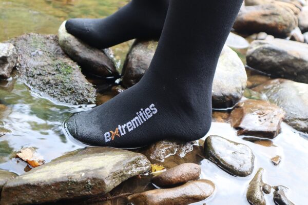 Close-up of a black sock branded 'extremities' on a foot standing on wet river stones with shallow water flowing around them.