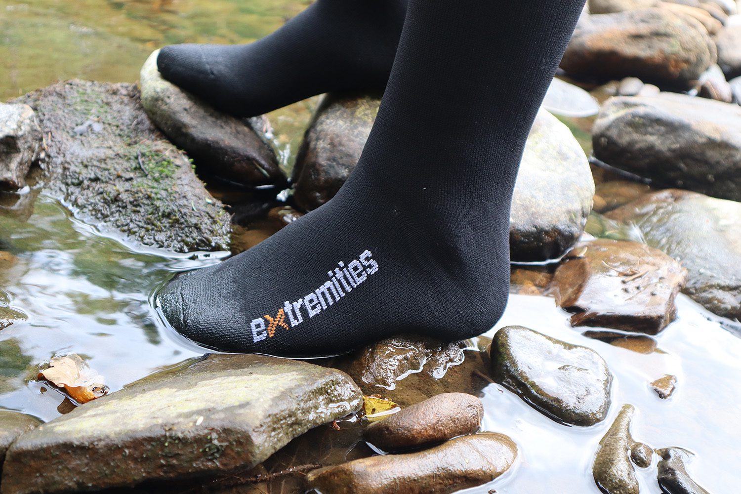 Close-up of a black sock branded 'extremities' on a foot standing on wet river stones with shallow water flowing around them.