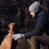 Igneous-and-Bakewell-scaled-2.jpg Man in a grey beanie and navy quilted jacket sits on a bench outdoors, reaching with a gloved hand to pet a brown short‑haired dog wearing a rope collar, with an old stone building in the background.
