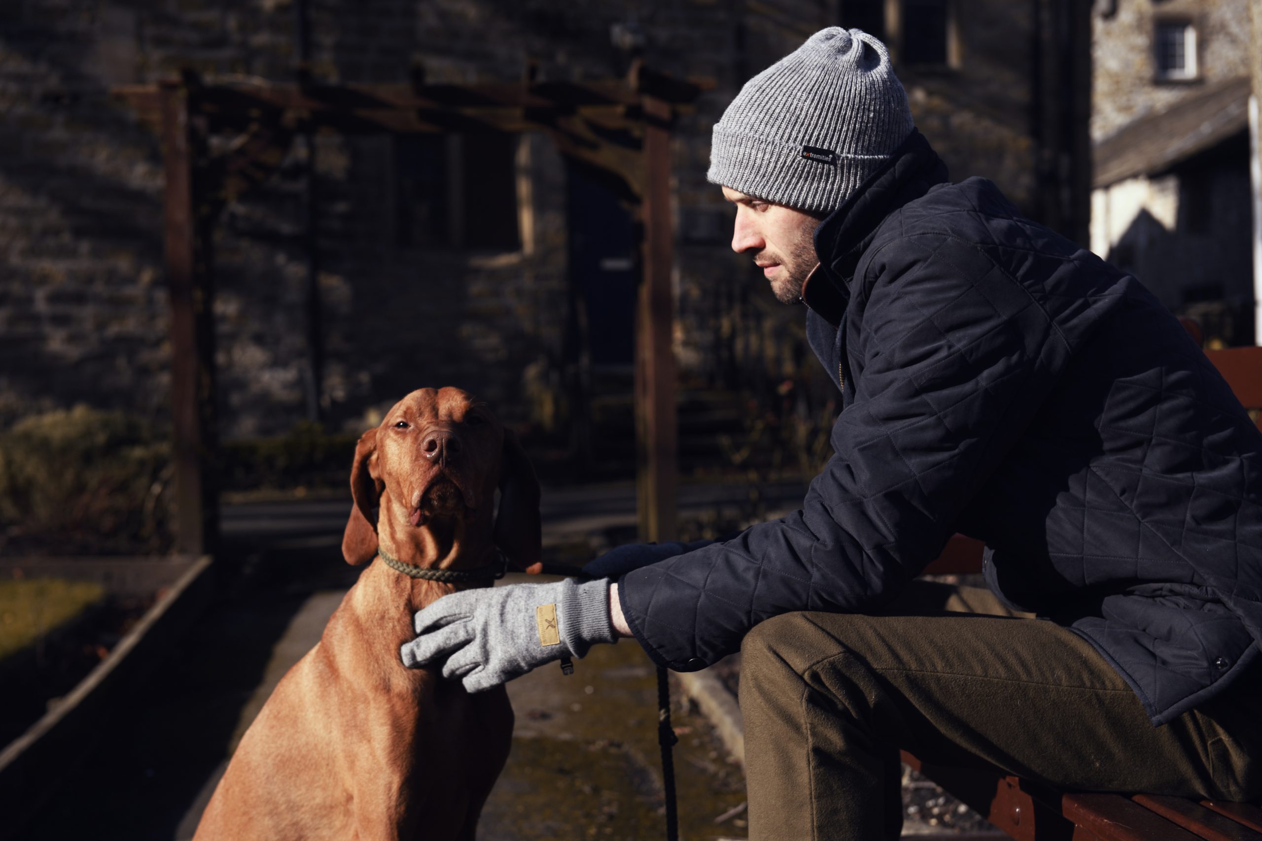 Igneous-and-Bakewell-scaled-2.jpg Man in a grey beanie and navy quilted jacket sits on a bench outdoors, reaching with a gloved hand to pet a brown short‑haired dog wearing a rope collar, with an old stone building in the background.