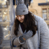 Young woman in a grey wool coat and matching pom‑pom knit hat leans forward to peer at a shop window, her long dark hair over one shoulder and a gloved hand near her chest, with warm display lights and her soft reflection in the glass behind her.