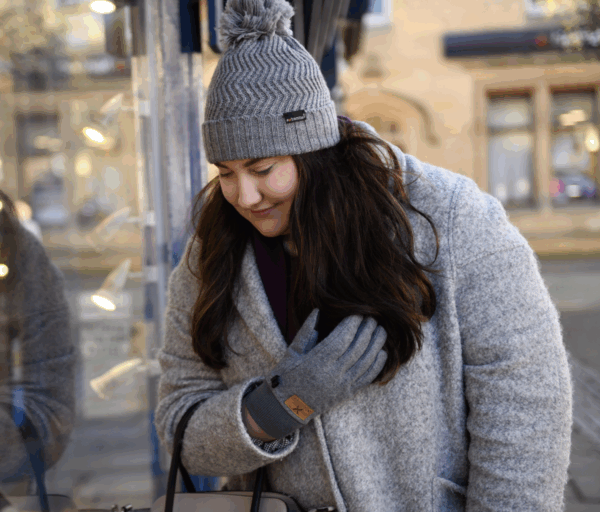Young woman in a grey wool coat and matching pom‑pom knit hat leans forward to peer at a shop window, her long dark hair over one shoulder and a gloved hand near her chest, with warm display lights and her soft reflection in the glass behind her.