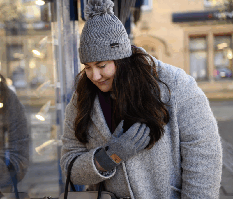 Igneous-and-castleton1-1.png Young woman in a grey wool coat and matching pom‑pom knit hat leans forward to peer at a shop window, her long dark hair over one shoulder and a gloved hand near her chest, with warm display lights and her soft reflection in the glass behind her.