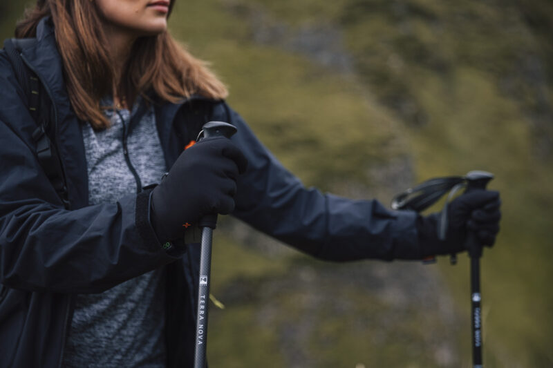 Insulated Contact Waterproof Power Liners (3) Close-up of a woman wearing gloves and a jacket, holding a hiking pole while exploring a mountainous landscape. The image captures the spirit of outdoor adventure and hiking gear essentials.