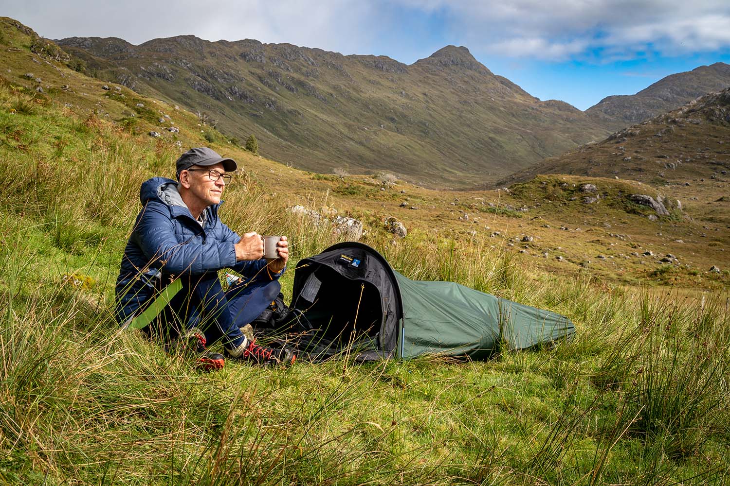 Jupiter-Lite-Bivi-10-LR-3.jpg Man wearing a blue insulated jacket and cap sits on grass beside a small green bivvy tent, holding a mug and gazing across a mountainous valley under a partly cloudy sky.