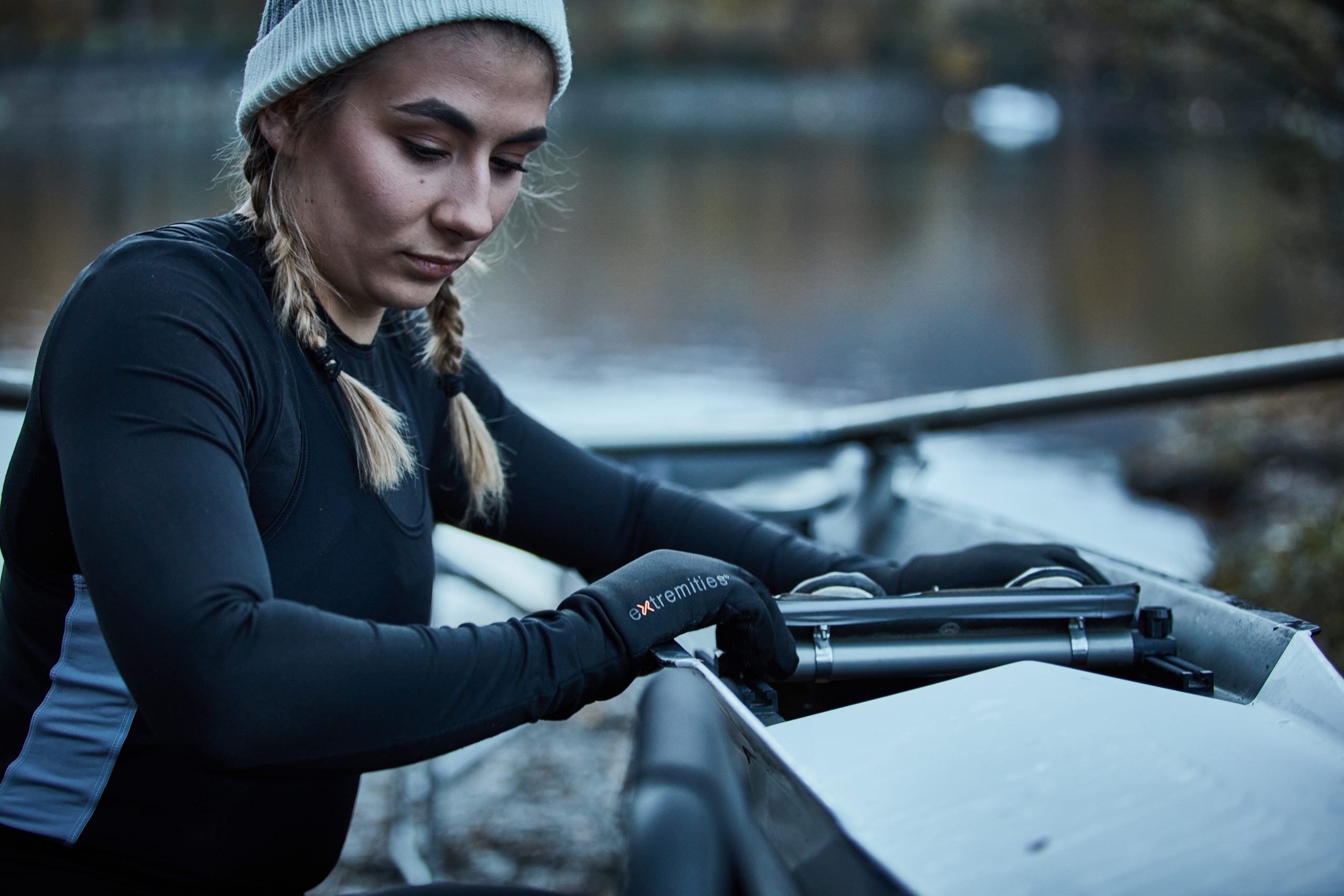 Young woman in a beanie and braided pigtails wearing a black thermal top and gloves adjusts the rigging on a rowing shell beside a calm river.