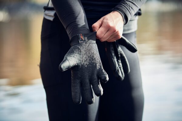 Close-up of a person pulling a textured black neoprene glove onto their hand while wearing a black wetsuit, with blurred water in the background.