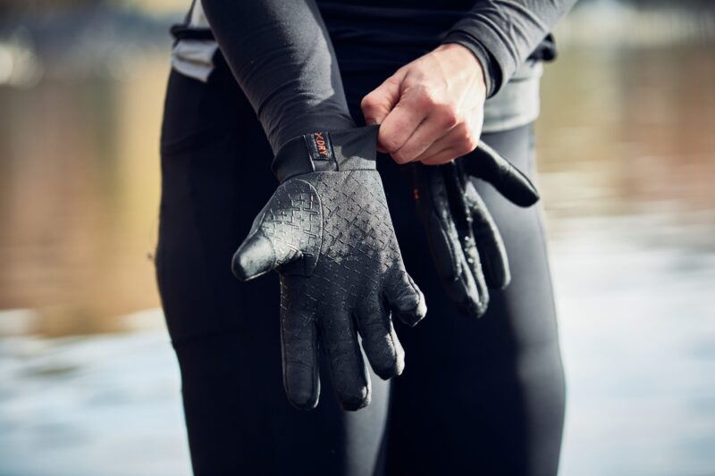 LIN-TERRANOVA-211109-1603-scaled-4.jpg Close-up of a person pulling a textured black neoprene glove onto their hand while wearing a black wetsuit, with blurred water in the background.