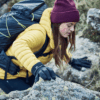 Woman wearing a maroon beanie, yellow puffer jacket and black gloves climbs over lichen-covered rocks with a large hiking backpack, reaching forward with one gloved hand on a rocky hillside.