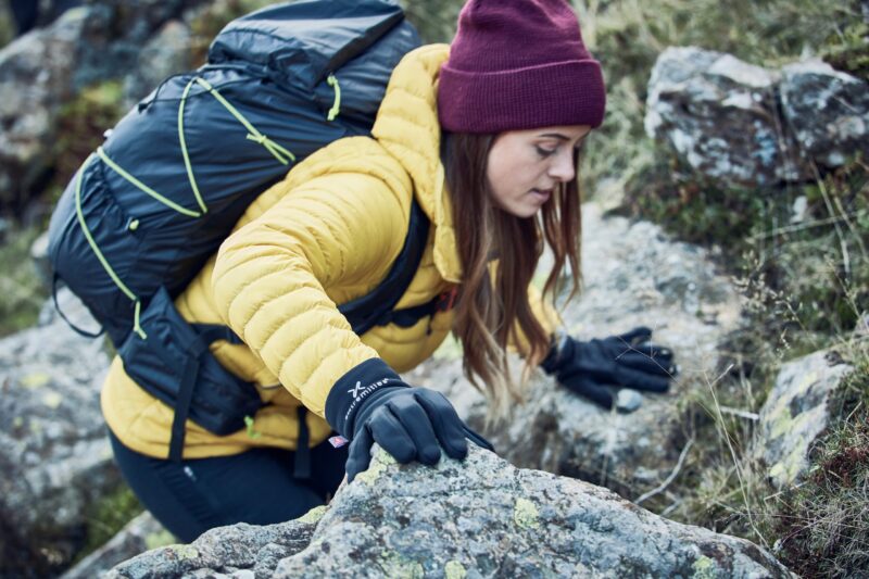 LIN-TERRANOVA-211109-2198-scaled-2.jpg A hiker in a yellow down jacket and maroon beanie, wearing black gloves and a dark backpack, climbs over lichen-covered rocks and reaches forward with a gloved hand.