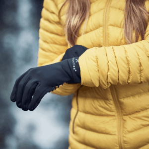 Close-up of a person in a yellow quilted jacket pulling on black thermal gloves against a blurred outdoor background.