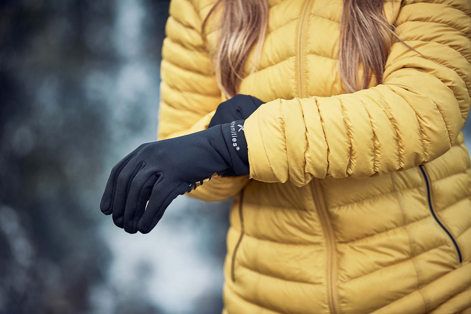 LIN-TERRANOVA-211109-2280-3.png Close-up of a person in a yellow quilted jacket pulling on black thermal gloves against a blurred outdoor background.