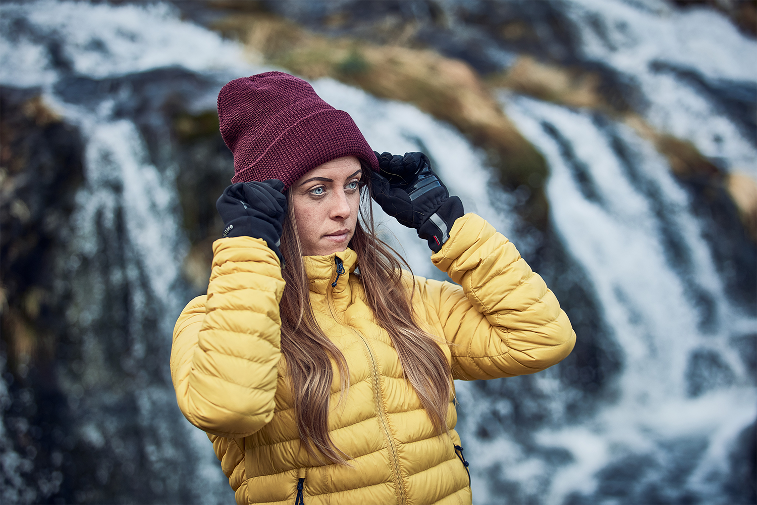 LIN-TERRANOVA-211109-2297-3.png Person in a yellow puffer jacket and black gloves adjusting a maroon beanie with long hair and a focused expression, standing in front of a blurred waterfall.