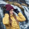 LIN-TERRANOVA-211109-2297-scaled-2.jpg Woman in a yellow quilted jacket adjusting a maroon beanie with black gloves while standing in front of a blurred cascading waterfall