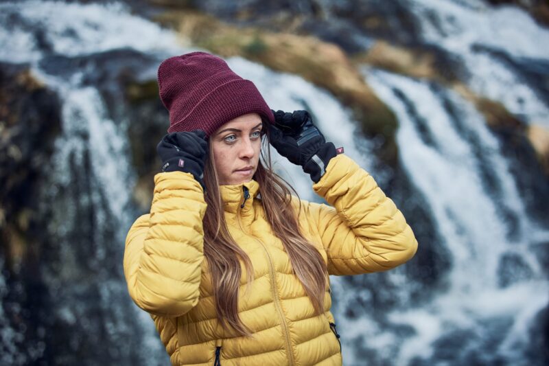 LIN-TERRANOVA-211109-2297-scaled-2.jpg Woman in a yellow quilted jacket adjusting a maroon beanie with black gloves while standing in front of a blurred cascading waterfall