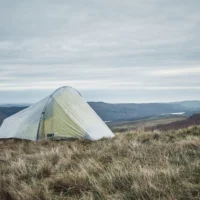 A pale green backpacking tent pitched on windswept grassy moorland overlooking rolling hills under a cloudy, overcast sky.