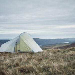 A pale green backpacking tent pitched on windswept grassy moorland overlooking rolling hills under a cloudy, overcast sky.