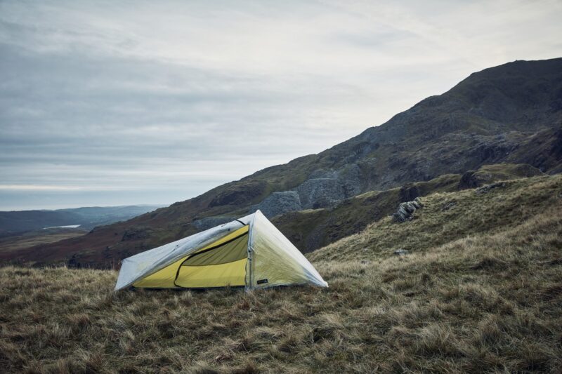 LIN-TERRANOVA-211109-2340-scaled-4.jpg A lightweight tent with a yellow inner and translucent rainfly pitched on a windswept grassy hillside, steep rocky slopes rising behind it under a pale cloudy sky.