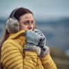 Woman in a yellow puffer jacket and fur earmuffs pressing grey, fur-trimmed mittens to her face while looking into a blurred cold outdoor landscape.