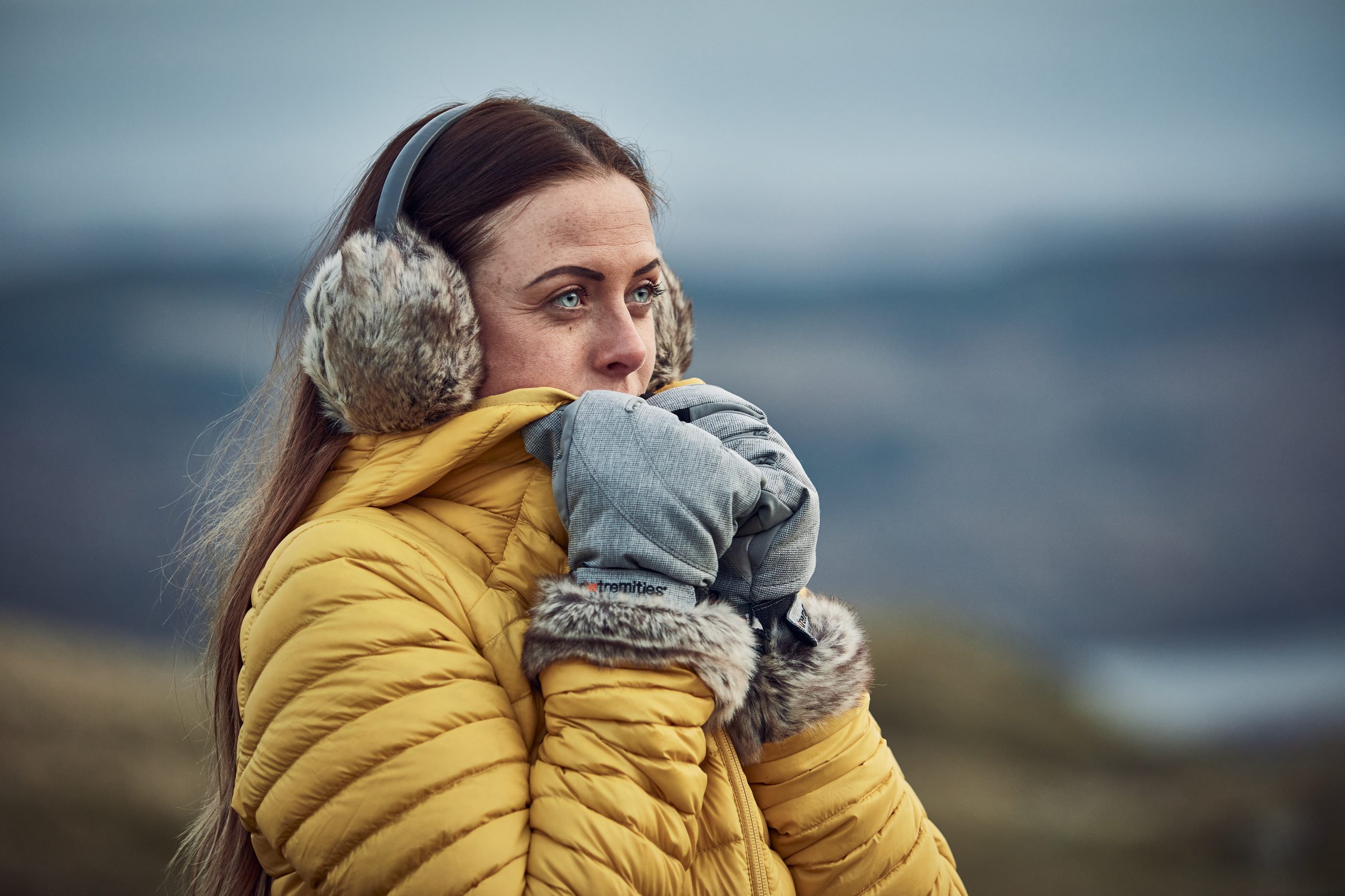 LIN-TERRANOVA-211109-2403-scaled-2.jpg Woman in a yellow puffer jacket and fur earmuffs pressing grey, fur-trimmed mittens to her face while looking into a blurred cold outdoor landscape.