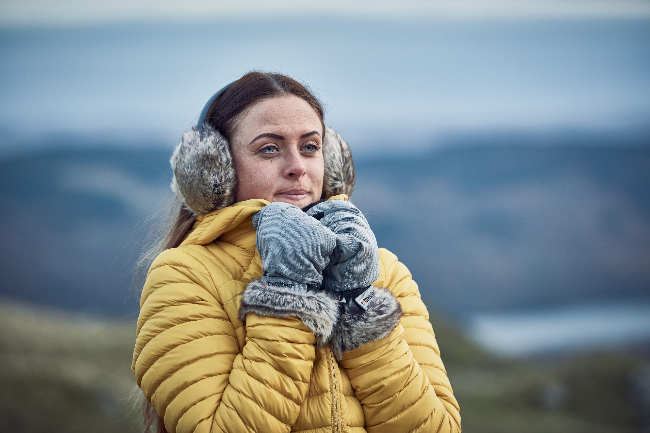 LIN-TERRANOVA-211109-2406-scaled-2.jpg Woman in a yellow quilted jacket wearing grey furry earmuffs and grey mittens with fur trim, holding her hands near her face and gazing into the distance against a blurred cold, hilly coastal background.