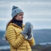 LIN-TERRANOVA-211109-2417-scaled-2.jpg Woman in a grey beanie and yellow puffer jacket with fur-lined grey mittens and a black neck gaiter, looking to the right against a soft-focus backdrop of hills and a cloudy sky.