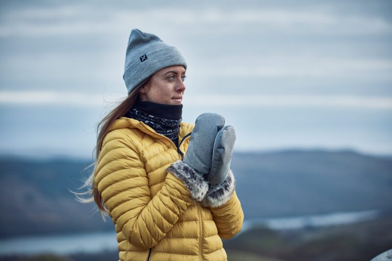 Woman in a grey beanie and yellow puffer jacket with fur-lined grey mittens and a black neck gaiter, looking to the right against a soft-focus backdrop of hills and a cloudy sky.