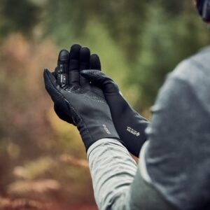 Hands of a person outdoors wearing black GORE‑TEX gloves held together as they adjust them, with a grey sleeve and a blurred green‑brown woodland background.