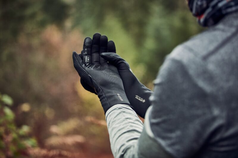 LIN-TERRANOVA-211109-3041-scaled-2.jpg Hands of a person outdoors wearing black GORE‑TEX gloves held together as they adjust them, with a grey sleeve and a blurred green‑brown woodland background.