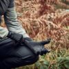 LIN-TERRANOVA-211109-3048-scaled-2.jpg Cropped view of a person kneeling in an autumnal fern-filled wood, pulling on textured black grip gloves while wearing a grey long-sleeve top and black trousers.