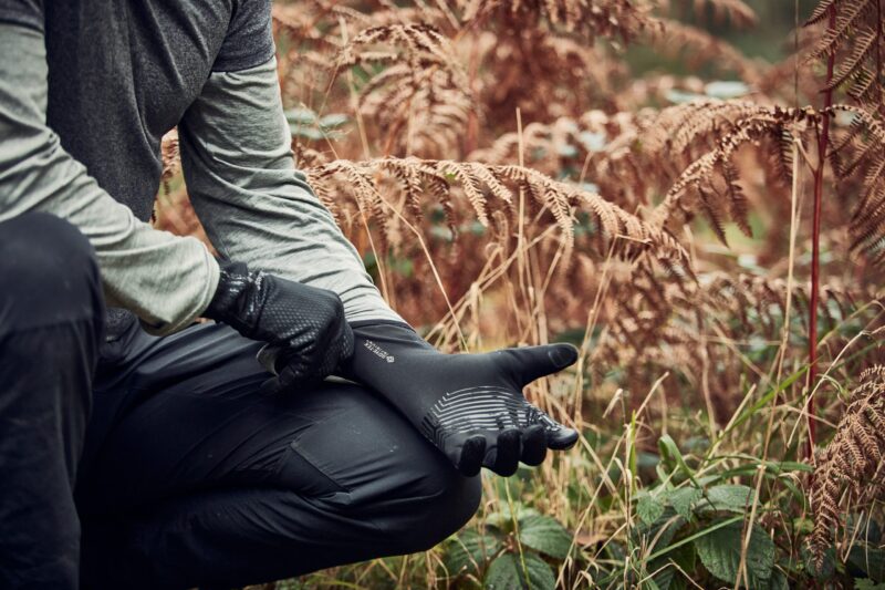 LIN-TERRANOVA-211109-3048-scaled-2.jpg Cropped view of a person kneeling in an autumnal fern-filled wood, pulling on textured black grip gloves while wearing a grey long-sleeve top and black trousers.